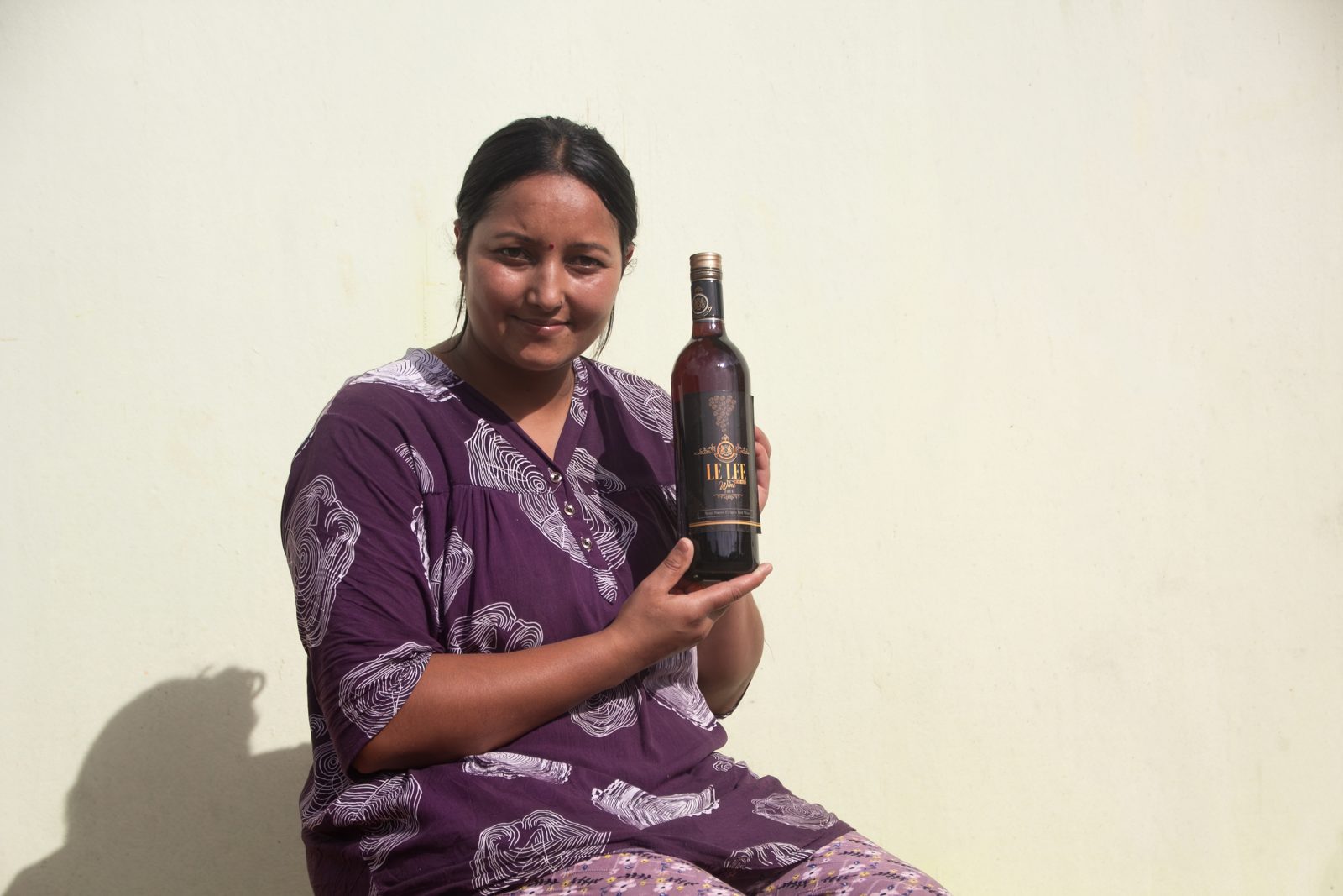 A maker of Le Le seated against a sunlit wall, holding a bottle of red